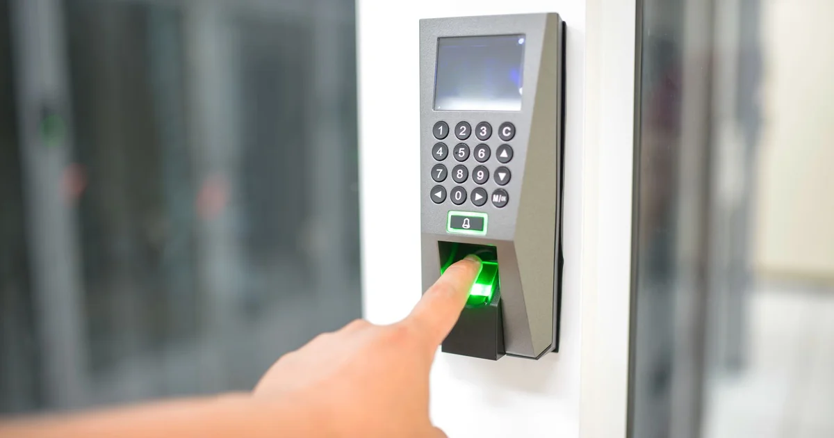Dedo sobre el lector de huella iluminado en verde de un terminal de control de acceso con teclado numérico y pantalla, junto a una puerta de vidrio