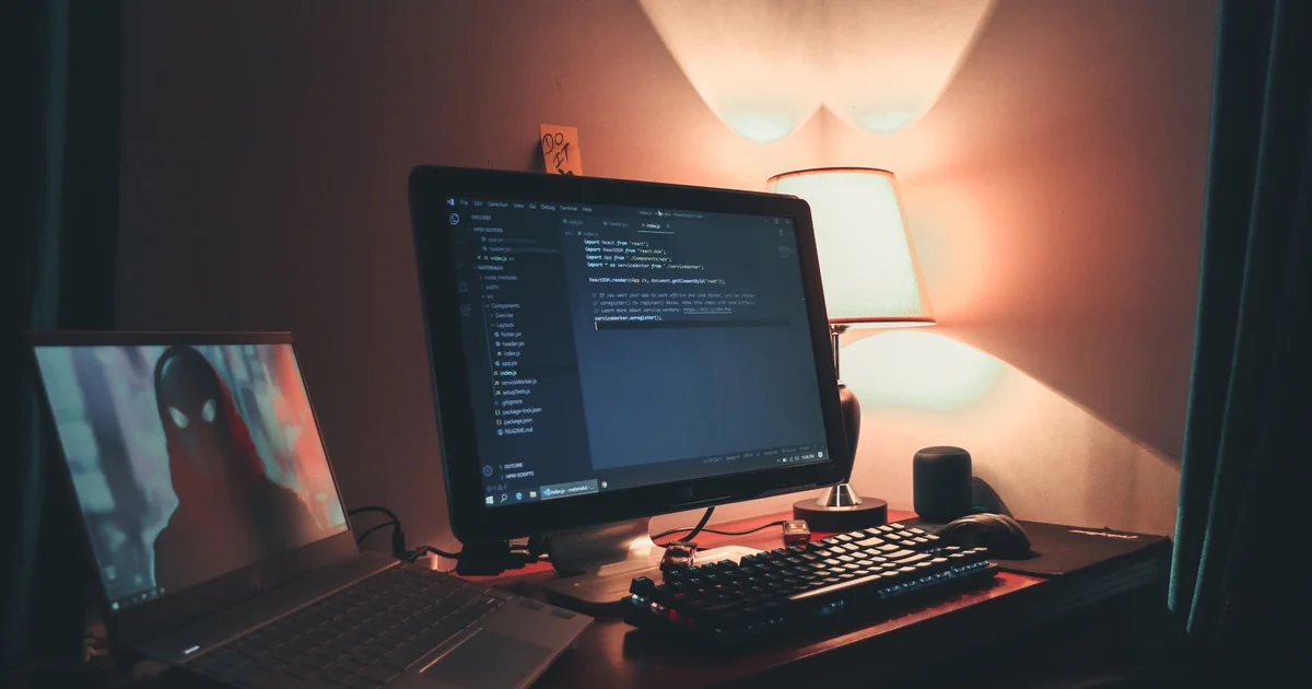 Escritorio en penumbra con un monitor que muestra código JavaScript en Visual Studio Code, un teclado iluminado, una lámpara encendida y, a la izquierda, una laptop con la imagen de una figura encapuchada