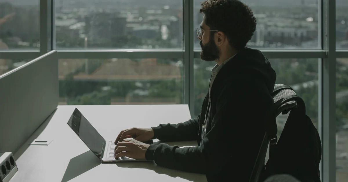 Hombre con barba y lentes, de sudadera negra, escribe en una laptop sobre un escritorio blanco de oficina frente a un ventanal con vista a la ciudad